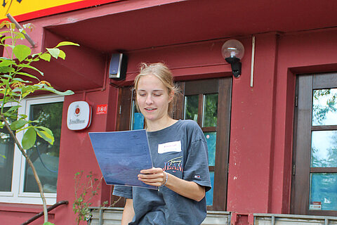 Apprentice recites her team's 8-liner, standing with her back to Bodega La Posta
